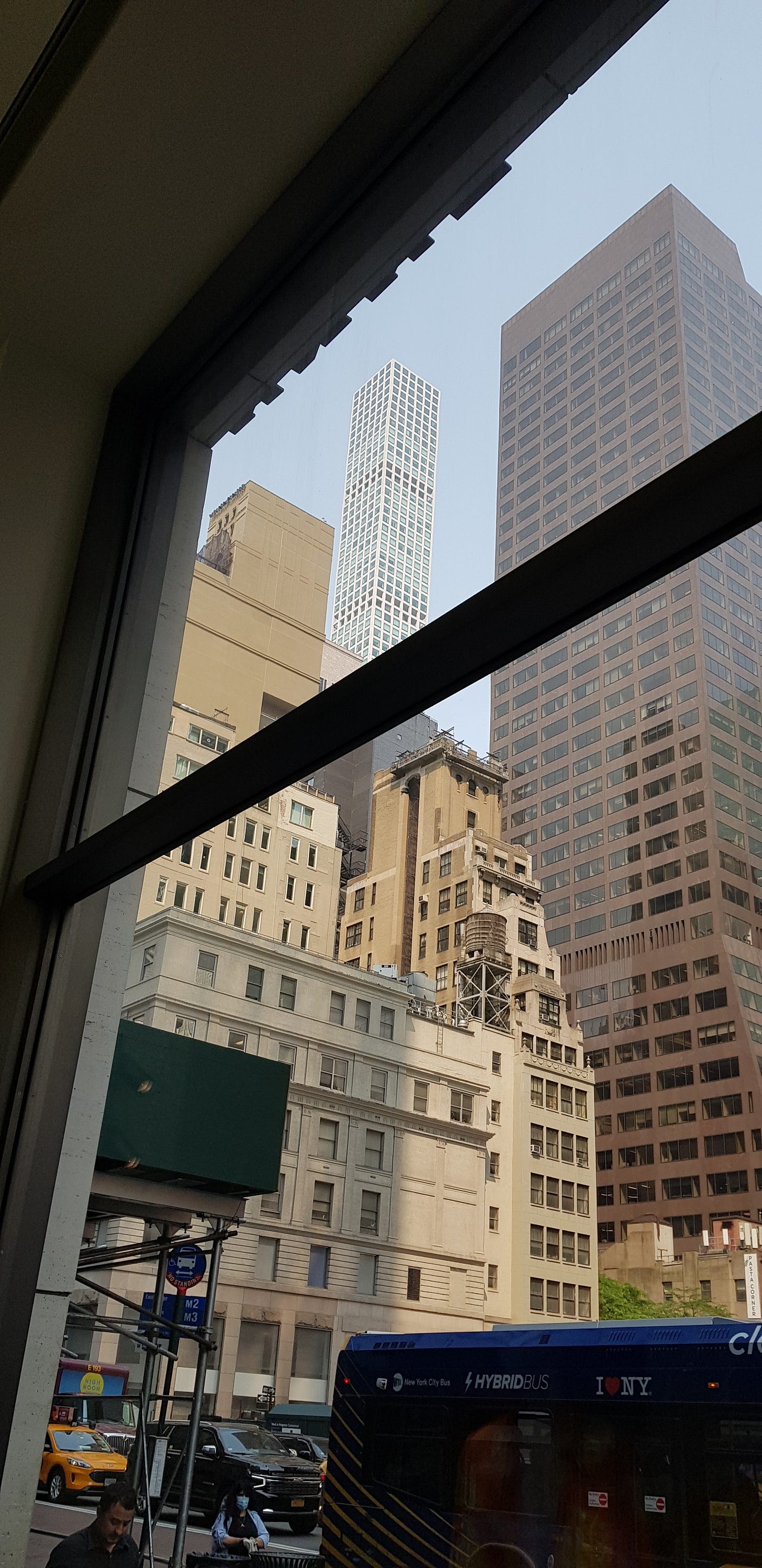 View from a small café window — a wooden water tank on a pre-war roof, a pencil-thin skyscraper behind it, a city bus passing below. View from a small café window — a wooden water tank on a pre-war roof, a pencil-thin skyscraper behind it, a city bus passing below.