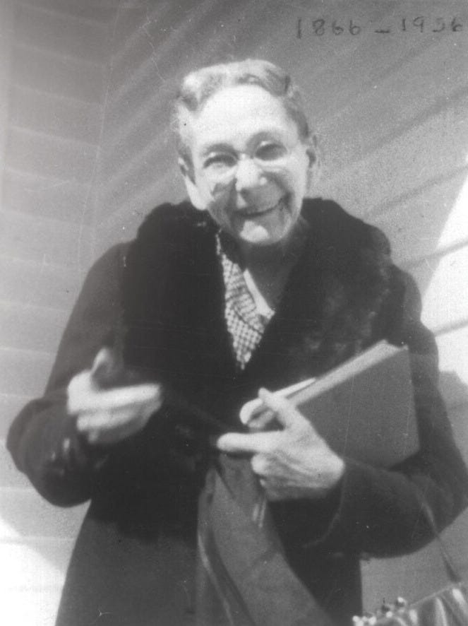 Elizabeth White in a black-and-white photo from around 1950. She is a puckish lady in her 80s, with a sweet smile and round wire-rimmed glasses. She wears a dark winter coat with a fur collar and holds two books in the crook of her left arm. A handle--possibly to an umbrella--is in her right hand.