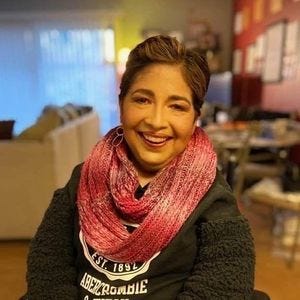 Michelle is smiling at the camera, wearing a knit red and pink scarf and a dark green sweater with fuzzy fleece sleeves.