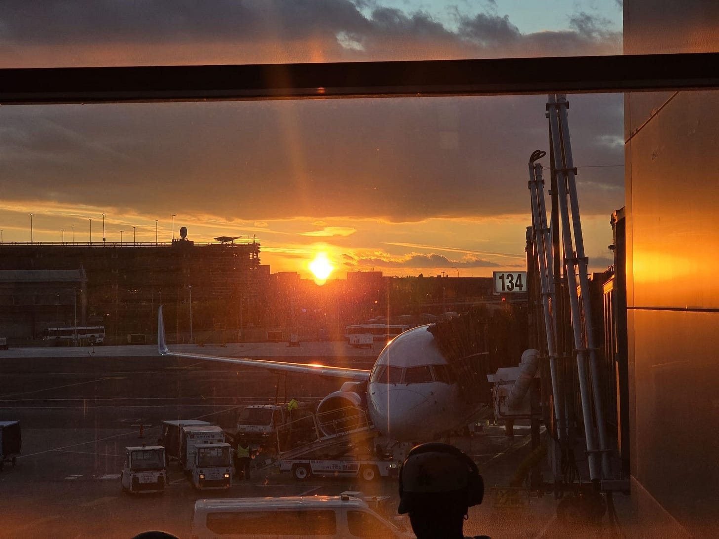 Photo out an airport window. A plane parked at the gate is visible with the sunrise behind it. Photo out an airport window. A plane parked at the gate is visible with the sunrise behind it.