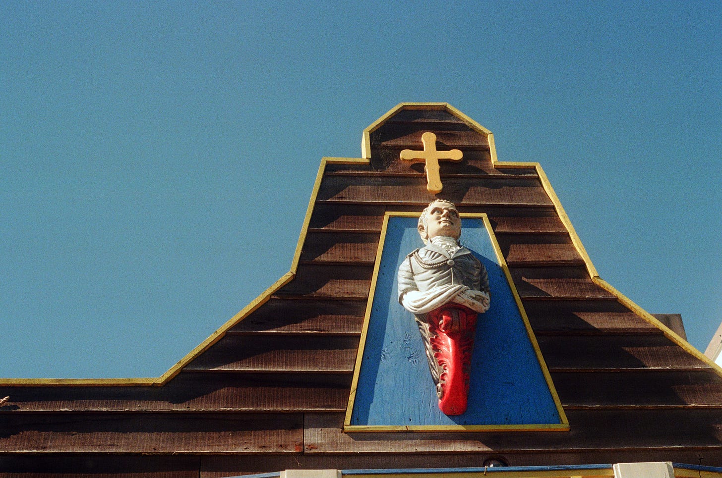 A film photograph of the top of a wooden building, where a manmade sculpture of a religious figure and cross have been hung. A bright blue sky can be seen in the background.