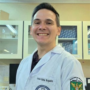 Na imagem, um homem sorri para a câmera dentro de um laboratório. Ele usa jaleco branco com nome bordado no peito e um emblema da FMRP no braço. Ao fundo, aparecem armários com portas de vidro, caixas, bancadas e equipamentos de laboratório.