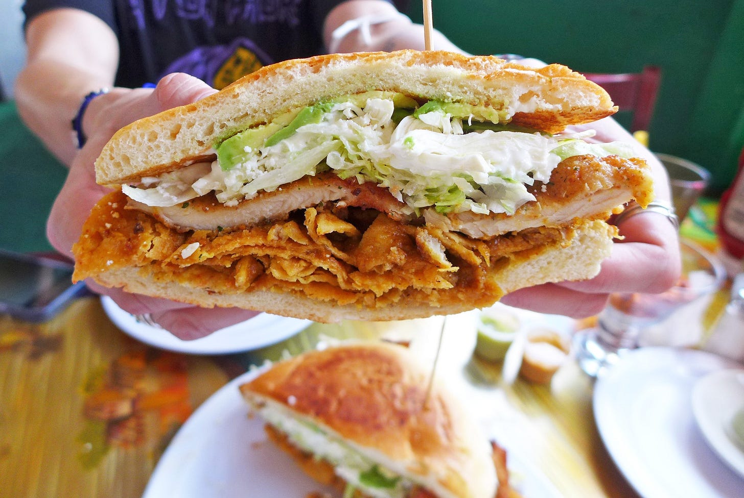 A sandwich held up by a pair of hands in which a fried cutlet and tortilla chips are visible.