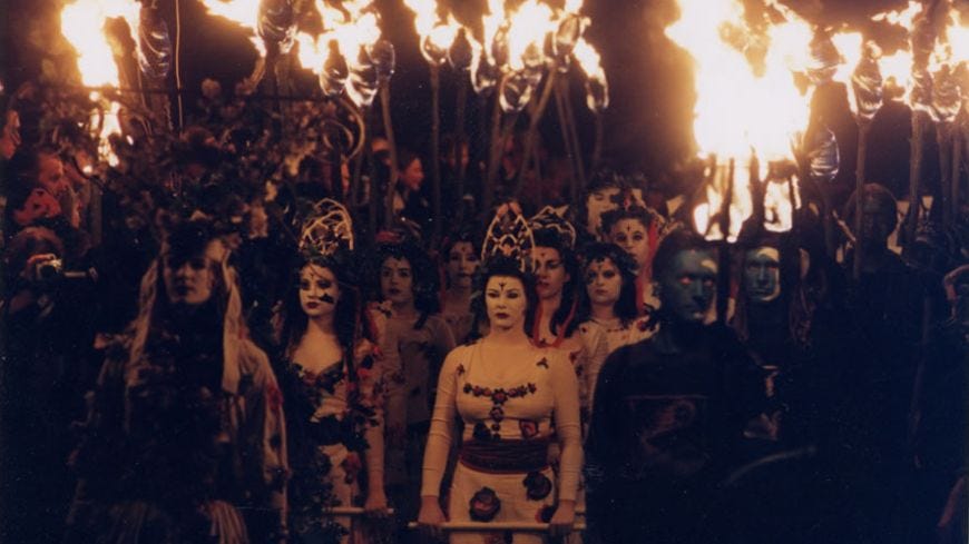 A dark photo of a procession of costumed people with face paint, crowns and torches. It is night and they are dressed like Celtic warriors and mythical druid like entities