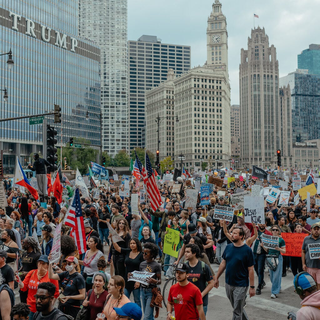 A group of people in an open area near a building with the word "Trump." Some people hold signs and American flags.
