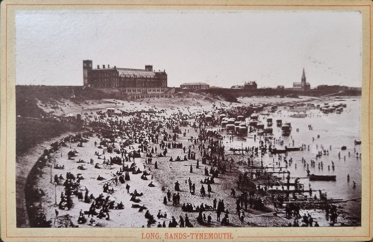 A historical black and white photograph of Long Sands Beach in Tynemouth, showing a large crowd of people enjoying the beach, with some walking along the shore and others sitting on the sand. Several carriages and structures are visible in the background.