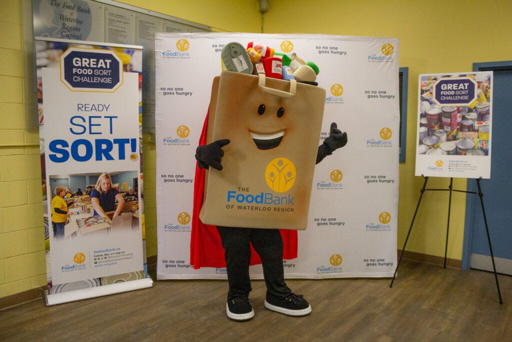 Food Bank mascot (a bag of groceries) stands in front of the Food Sort Challenge banner