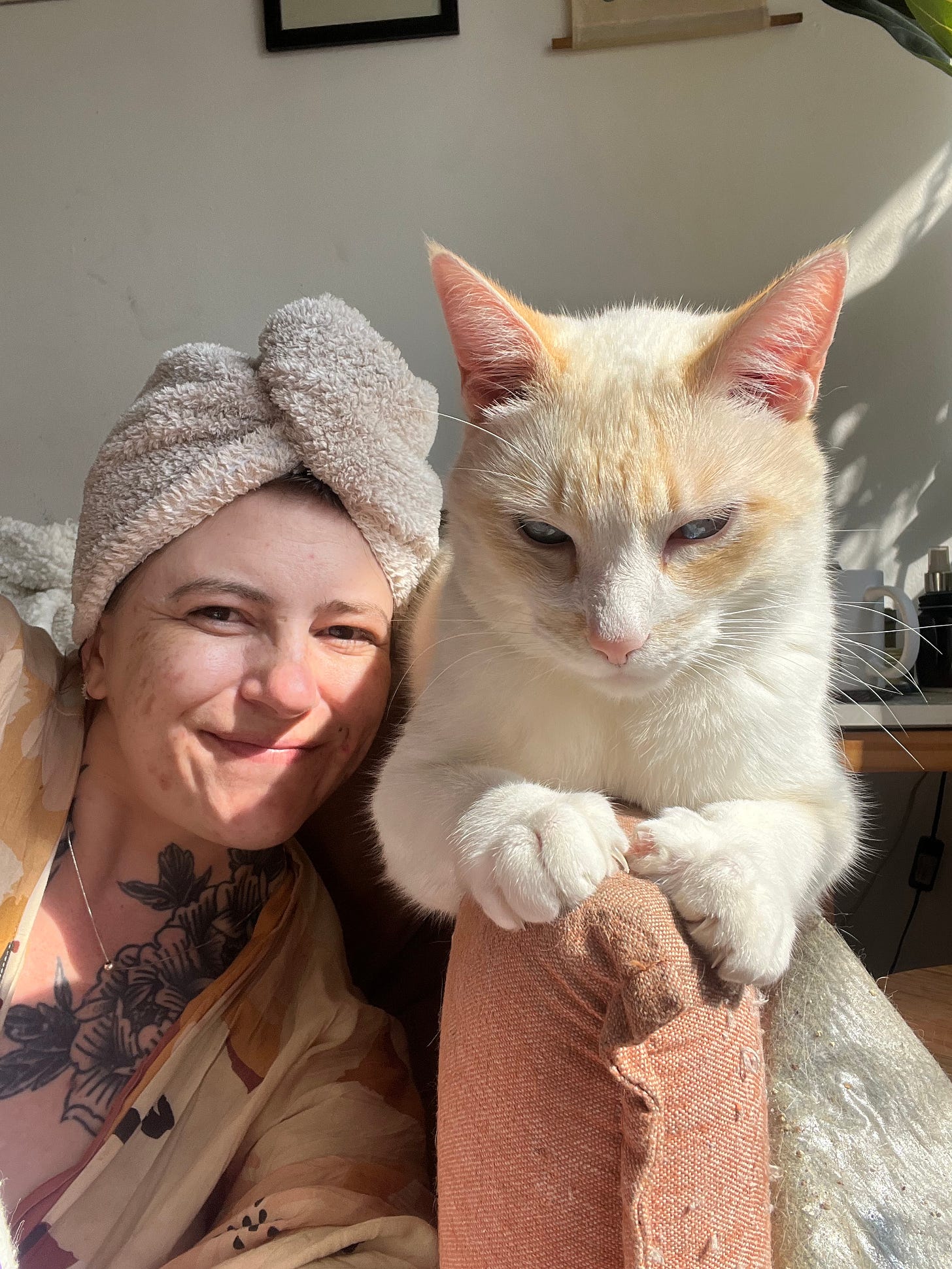 Selfie minha, mulher branca, toalha de banho enrolada na cabeça, com meu gato branco Bartolomeu, que está deitado no braço do sofá com os olhos semicerrados e as patinhas da frente agarrando o sofá. 