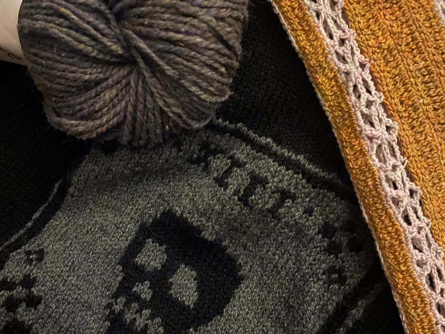 A pile of projects: a black and grey colorwork sweater depicting flowers and a skull; a grey/purple skein of Peace Fleece in colorway Sea Smoke; a gold and pink crocheted button up shirt destined for warmer months