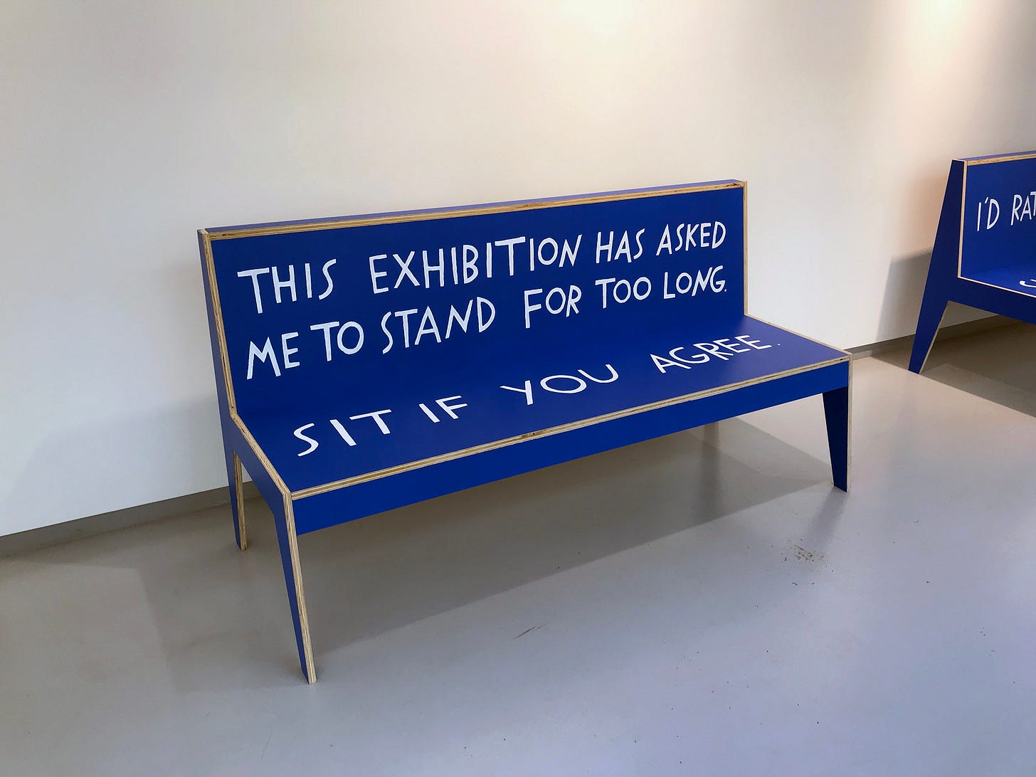 Una panchina in legno colora di azzurro presenta una scritta bianca sullo schienale e sulla seduta: "This exhibition has asked me to stand for too long. Sit if you agree." Una panchina in legno colora di azzurro presenta una scritta bianca sullo schienale e sulla seduta: "This exhibition has asked me to stand for too long. Sit if you agree."