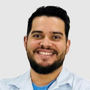 Homem branco, cabelos e olhos castanhos, usa jaleco branco e camiseta azul.