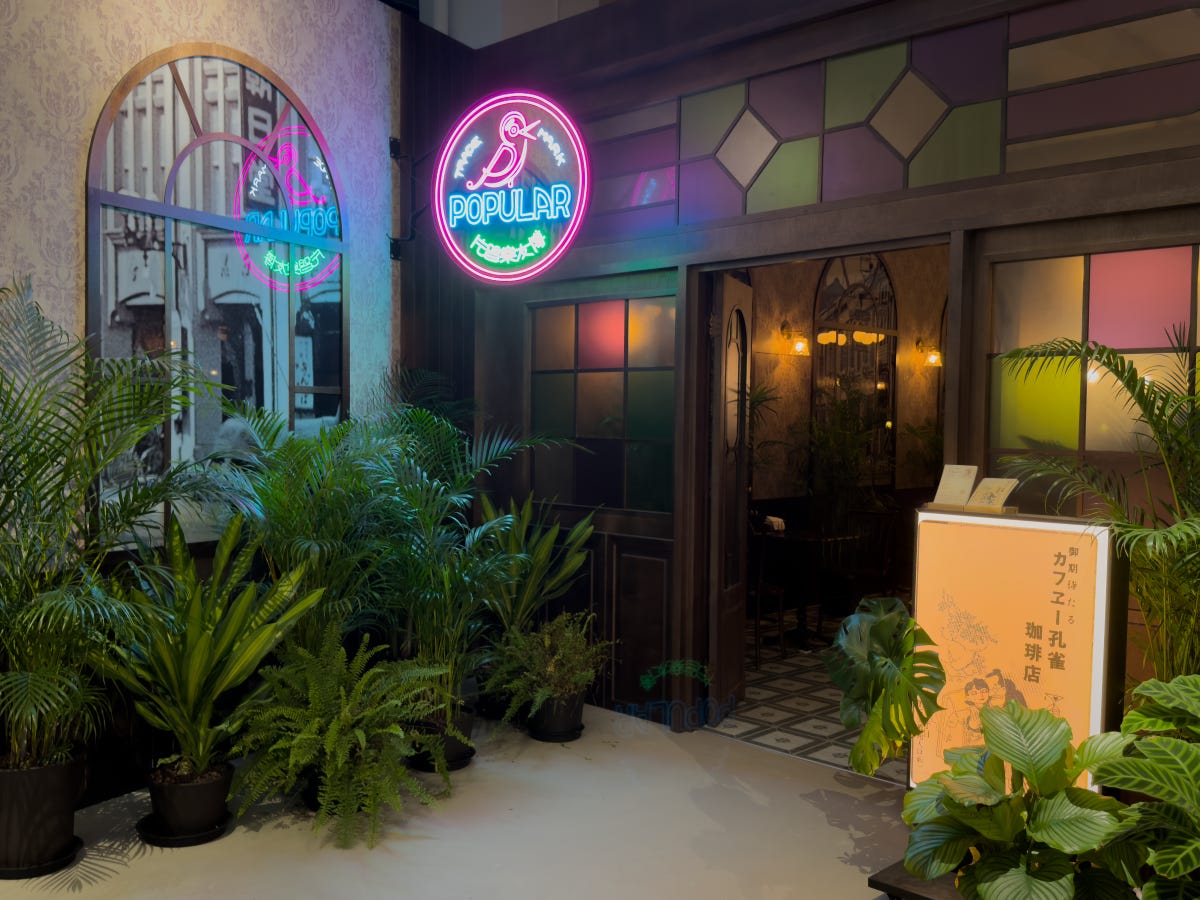 A pink, blue, and green neon sign featuring a bird and the word "popular" sits above the wood and stained-glass door to a cafe