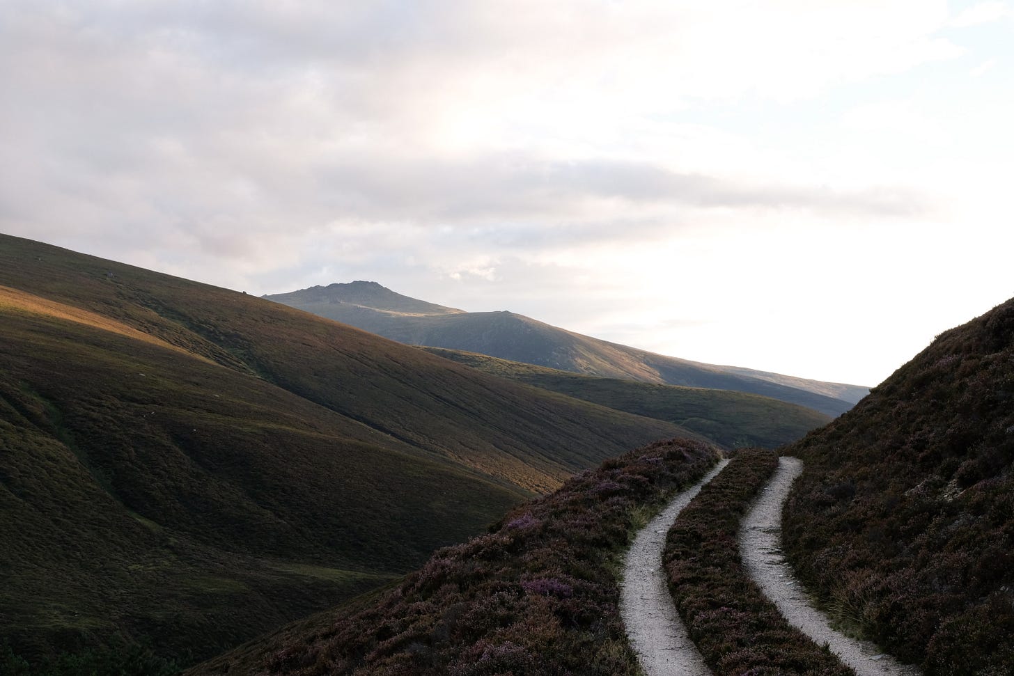 A narrow path lined with purple heather curves over the edge of a Highland hill, disappearing into a wide valley below. Dark green and purple heathers cover the hillsides, lit by swathes of golden sunlight breaking through moving clouds.