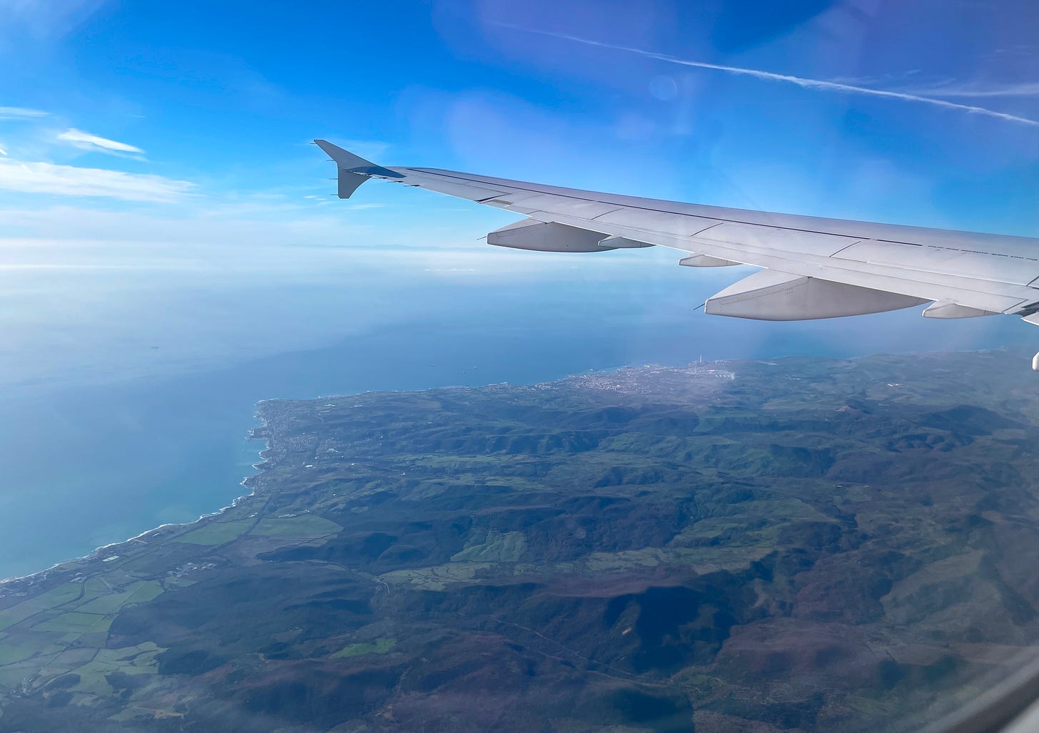 The view from a plane window, looking out over green land and a blue ocean. The wing of the plane is seen on the upper right side of the frame.