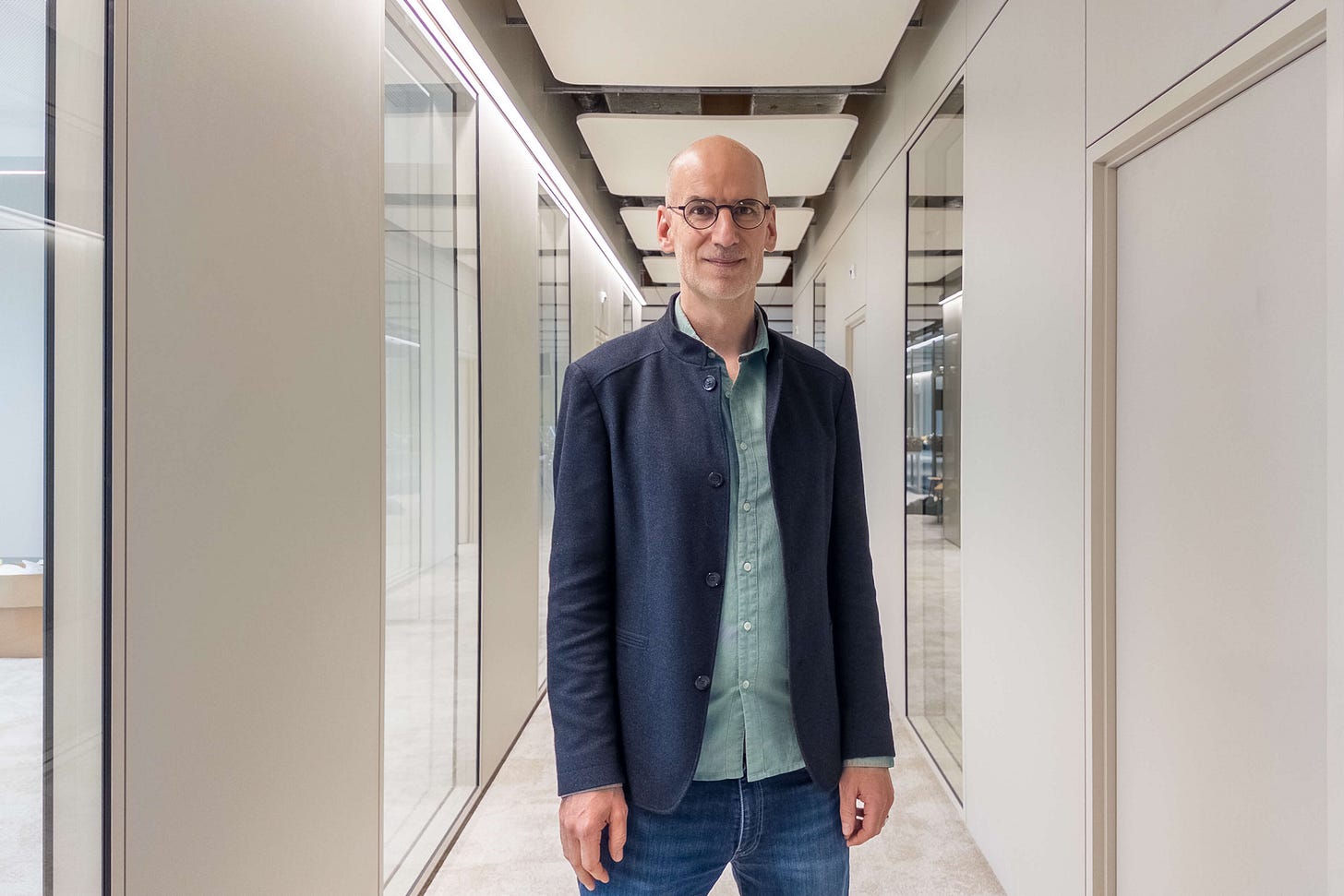 Balz Strasser, Co-Geschäftsführer von Bio Suisse, mit Glatze und runder Brille steht in einem hellen, modernen Flur zwischen Glastrennwänden und Türen. Er trägt ein hellgrünes Hemd, Jeans und ein dunkelblaues Sakko.