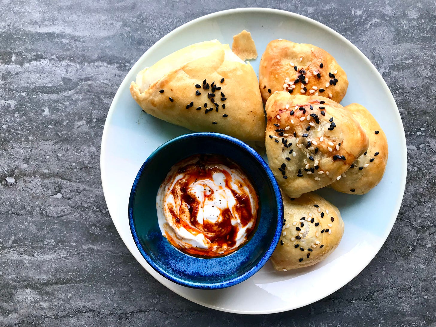 A plate of Uzbeck Beef Samsa, topped with black and white sesame seeds. They are golden and one (the large one, made with the flip and fold method, has s flake that's fallen off. The others are made with the Swiss roll method, and you can see a bit of the swirl on one. Next to the samsas is a blue bow with a swirly red and white dipping sauce made with sour cream and chilli crisp, with a sprinkling of black pepper. 