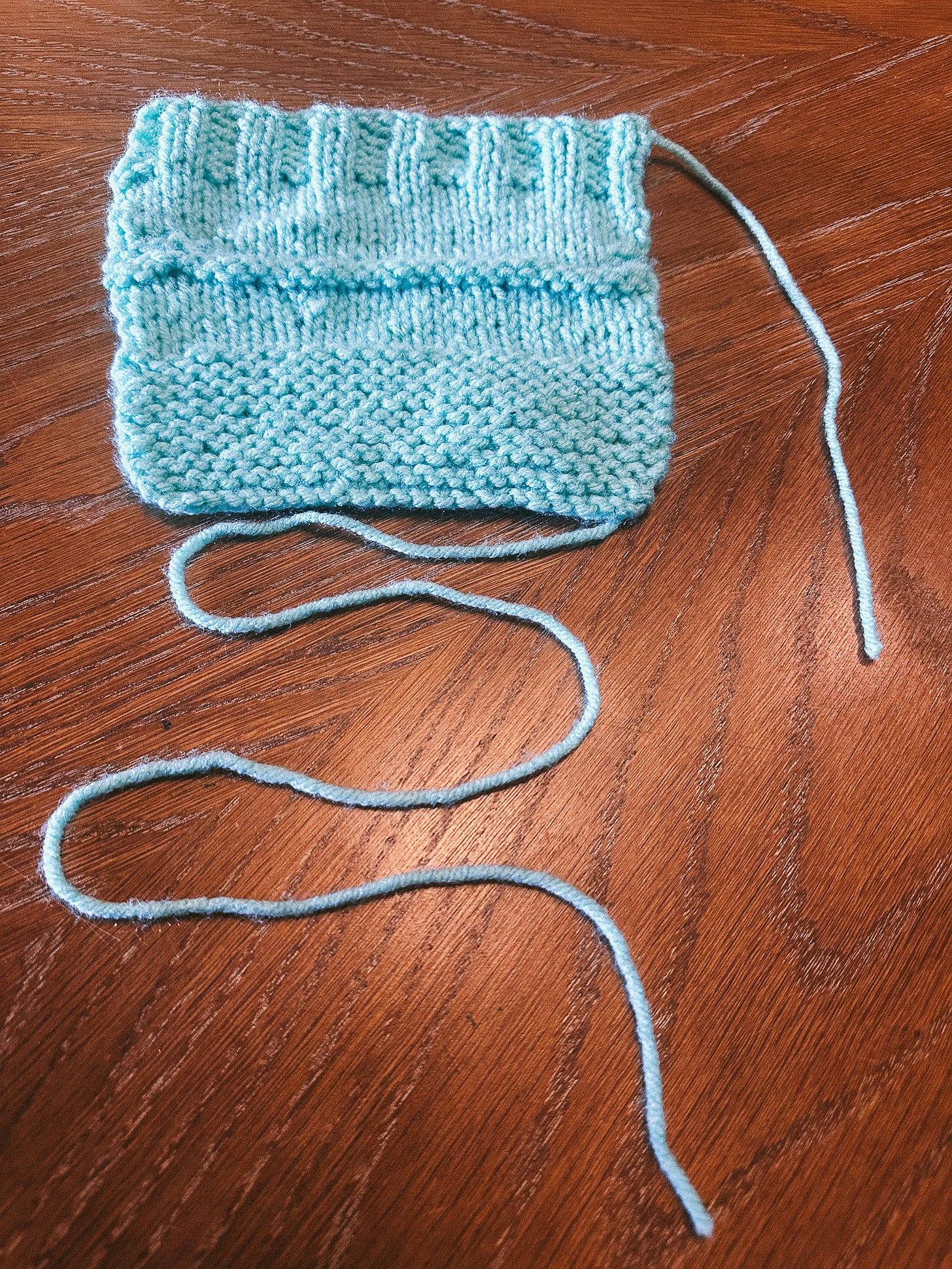 a light blue square of various knit stitches, a long tail of yarn curling beneath it a light blue square of various knit stitches, a long tail of yarn curling beneath it