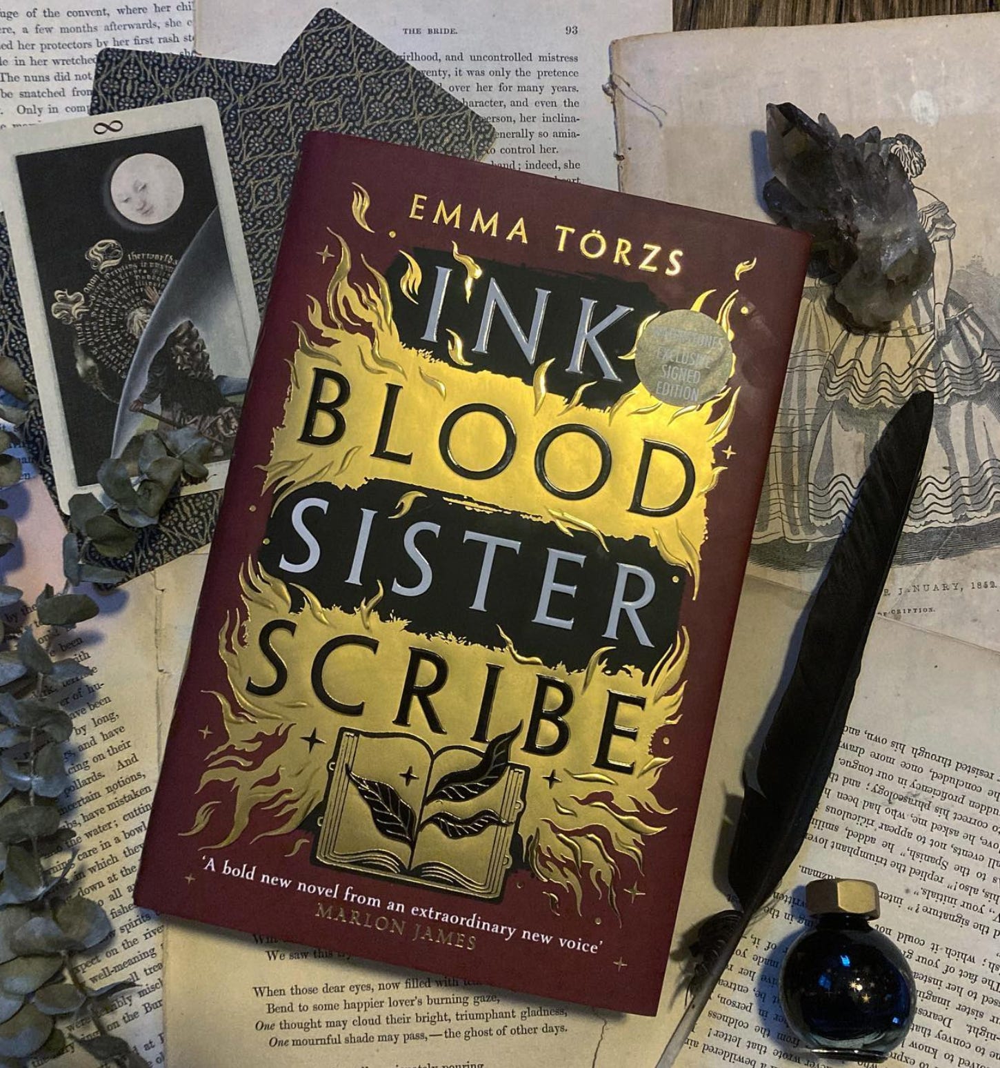A flat lay of Ink Blood Sister Scribe on a background of old book pages with tarot cards, dried eucalyptus, a black feather and a circular bottle of dark blue ink.