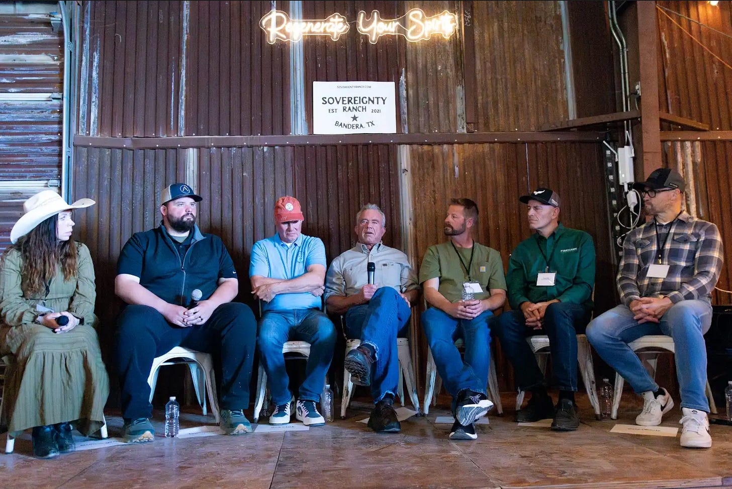 Panel of seven speakers including RFK Jr. seated in a barn beneath a neon "Regenerate Your Spirit" sign.