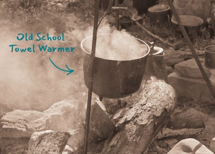 A black and white photo of a campfire with a cast iron pot boiling. There is an arrow pointing to the pot that says "old school towel warmer"