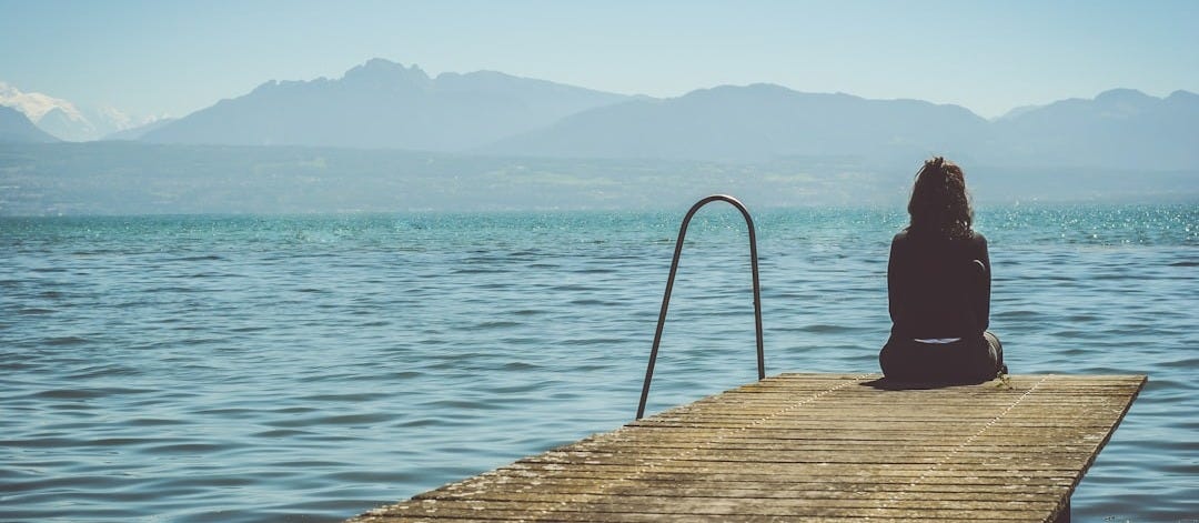 a woman sits on the end of a dock during daytime staring across a lake a woman sits on the end of a dock during daytime staring across a lake