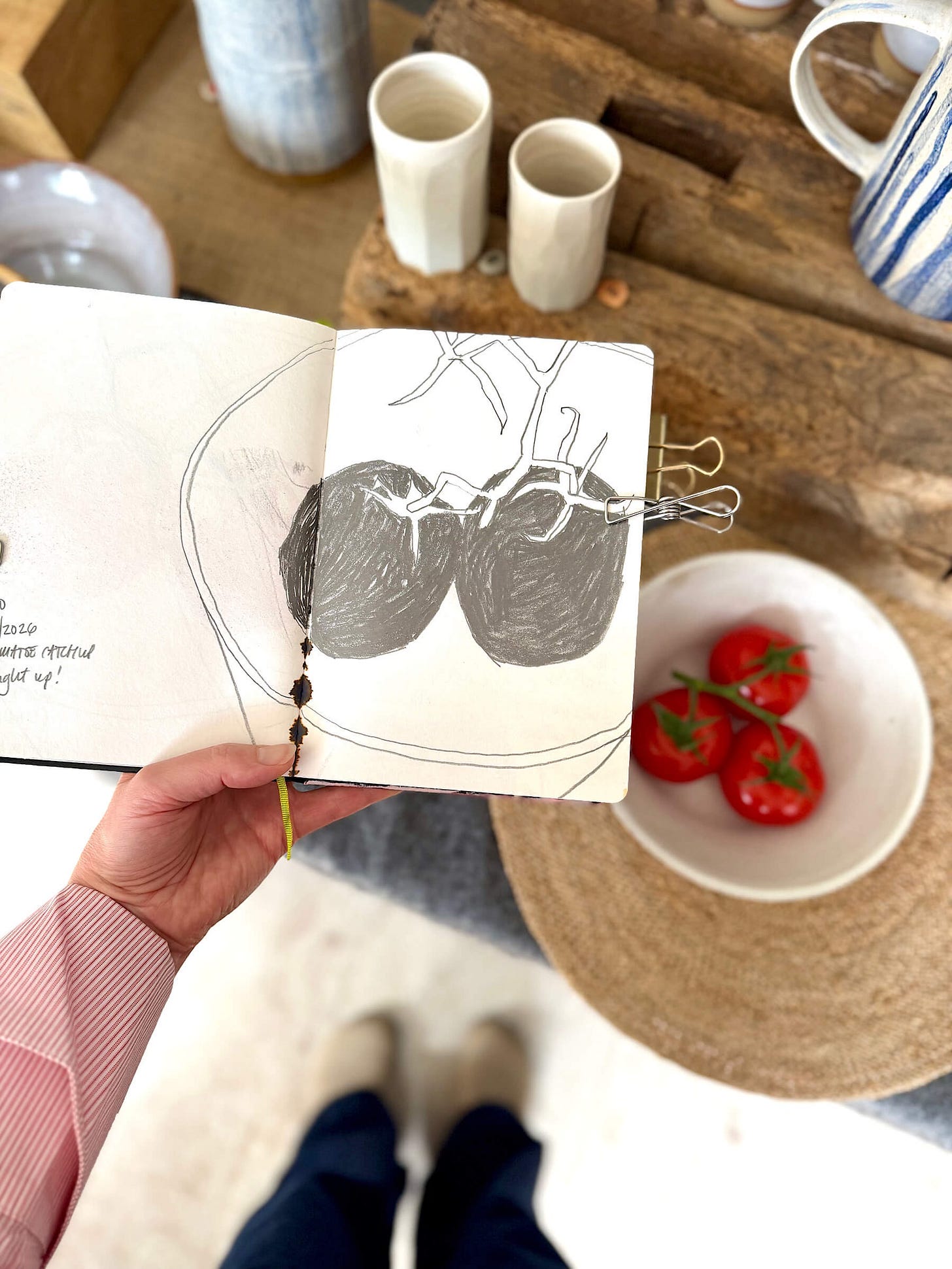 An overhead, first-person view of an artist holding an open sketchbook in a bright studio. The right-hand page displays a raw, initial graphite drawing of two tomatoes on a vine, with the left page containing partially legible text. Next to the sketch on a round placemat is a small ceramic bowl containing the real vine tomatoes that inspired the study. The soft-focus background shows ceramic vessels and an antique wooden block. An overhead, first-person view of an artist holding an open sketchbook in a bright studio. The right-hand page displays a raw, initial graphite drawing of two tomatoes on a vine, with the left page containing partially legible text. Next to the sketch on a round placemat is a small ceramic bowl containing the real vine tomatoes that inspired the study. The soft-focus background shows ceramic vessels and an antique wooden block.