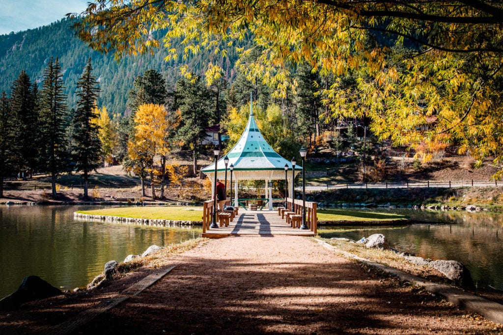 In Green Mountain falls- a gazebo stands on a small island surrounded by mountains covered with pine trees.