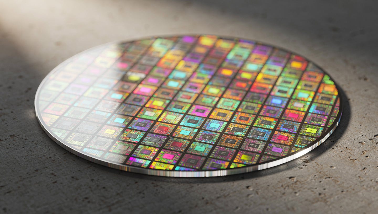 Realistic macro photograph of a 300mm silicon wafer with a grid of chips showing iridescent rainbow reflections. It rests on a warm grey brushed concrete surface, soft raking side light, wafer edge tack sharp, blurred background, no markings or text on the wafer, industrial editorial photography, high resolution.