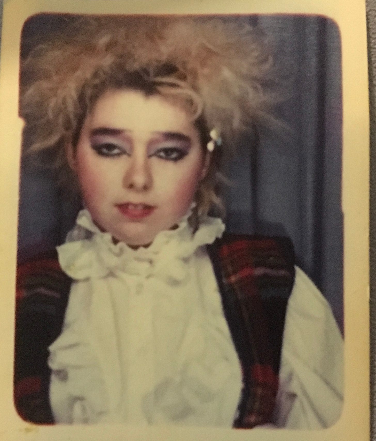 Young Penny with punky blonde wild hair, and heavy punky make up. She wears a high white collar frilly blouse and a tartan waistcoat. This photo is a head and shoulders shot with a blue curtain background. Penny is staring at the camera solemnly.