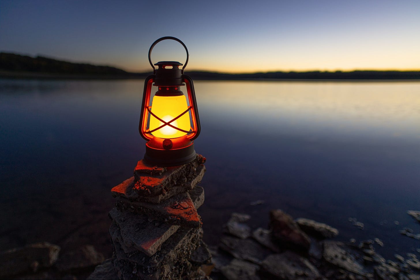 Image of a lit lantern on some rocks overlooking a body of water at dusk.