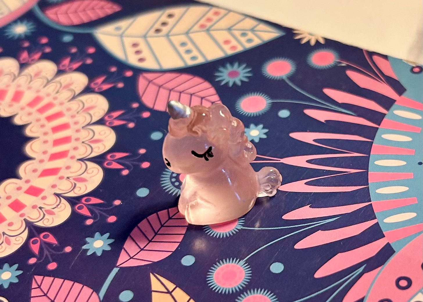 close up of a tiny pink unicorn figurine resting atop a notebook with a blue, pink and white retro floral pattern
