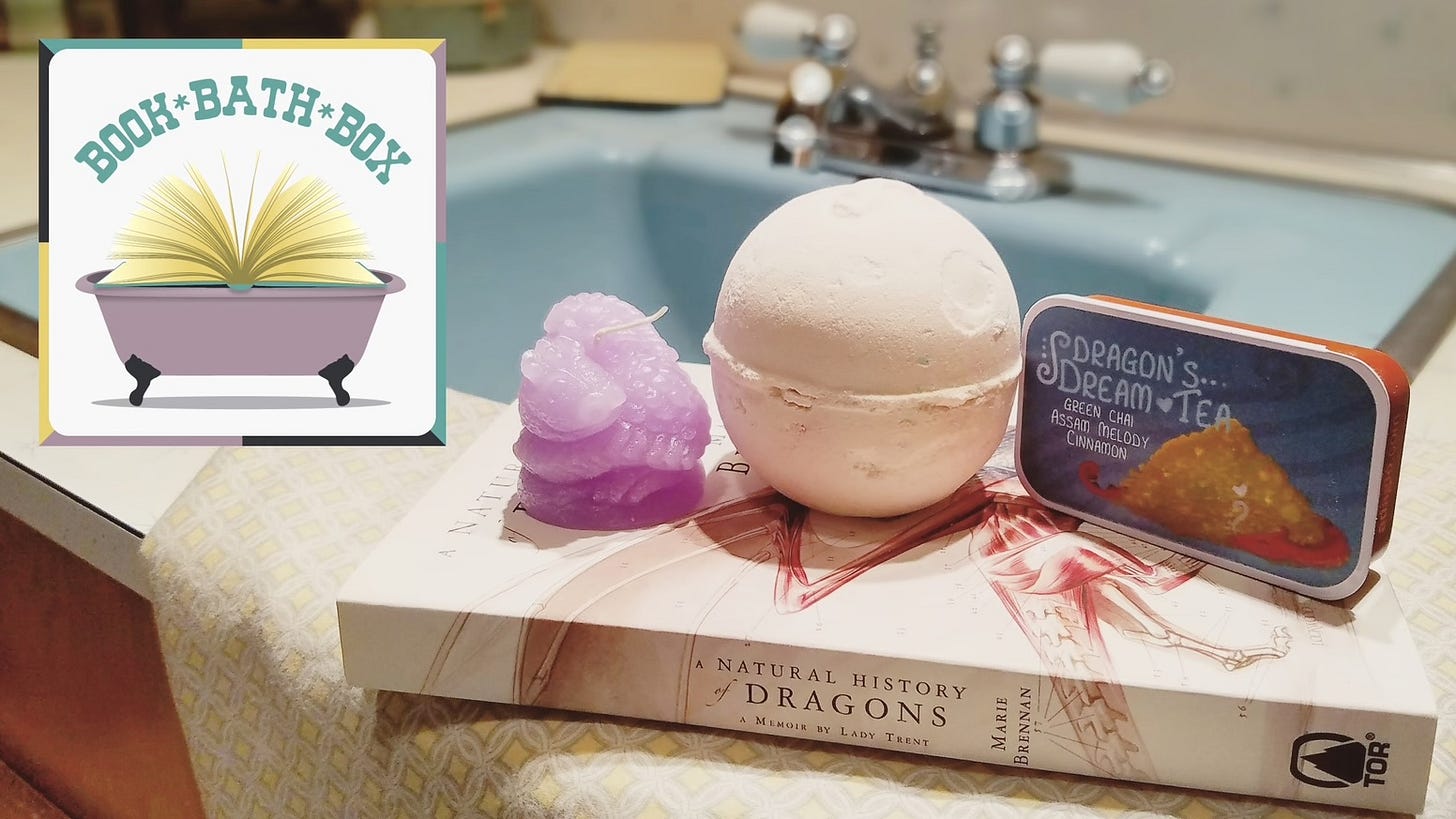 A cozy bath-time book box setup featuring the book A Natural History of Dragons by Marie Brennan placed beside a purple dragon-shaped candle, a large white bath bomb, and a tin of “Dragon’s Dream Tea” labeled as green chai, Assam melody, and cinnamon. In the background is a vintage-style blue bathtub with silver fixtures. An inset logo in the top left corner reads “Book * Bath * Box” with an open book inside a clawfoot tub, suggesting a themed self-care and reading subscription box experience. A cozy bath-time book box setup featuring the book A Natural History of Dragons by Marie Brennan placed beside a purple dragon-shaped candle, a large white bath bomb, and a tin of “Dragon’s Dream Tea” labeled as green chai, Assam melody, and cinnamon. In the background is a vintage-style blue bathtub with silver fixtures. An inset logo in the top left corner reads “Book * Bath * Box” with an open book inside a clawfoot tub, suggesting a themed self-care and reading subscription box experience.