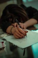 A woman with her head on a desk, pen in hand on a sheet of scribbled-over paper.