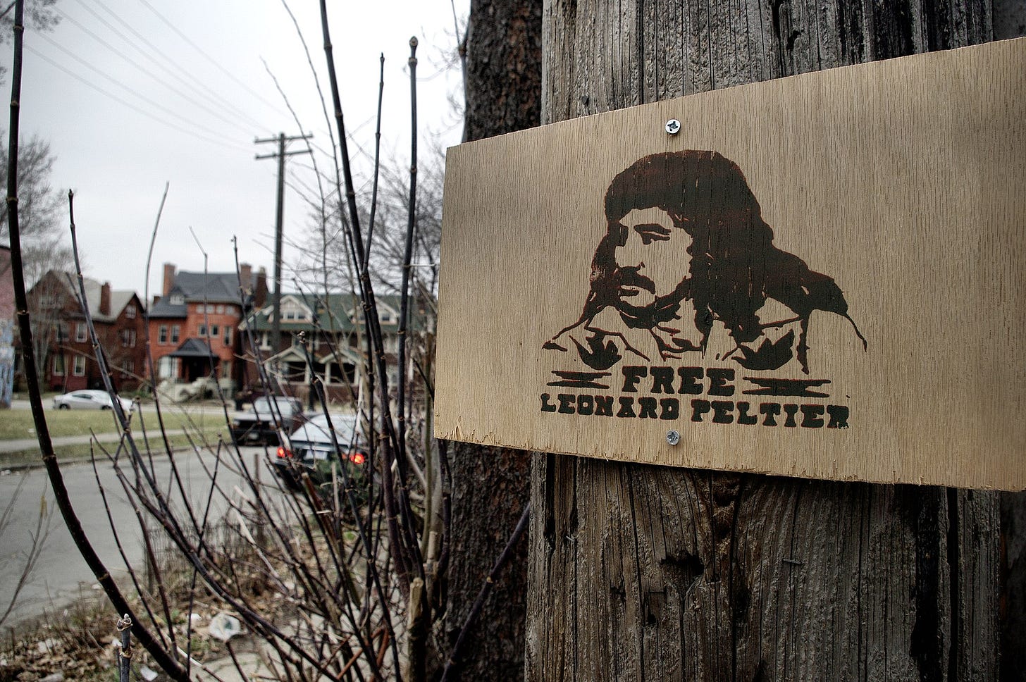 "Free Leonard Peltier" sign on a telephone pole. Illustration of Peltier with long hair and mustache; a street with parked cars is off to the side/in the background "Free Leonard Peltier" sign on a telephone pole. Illustration of Peltier with long hair and mustache; a street with parked cars is off to the side/in the background