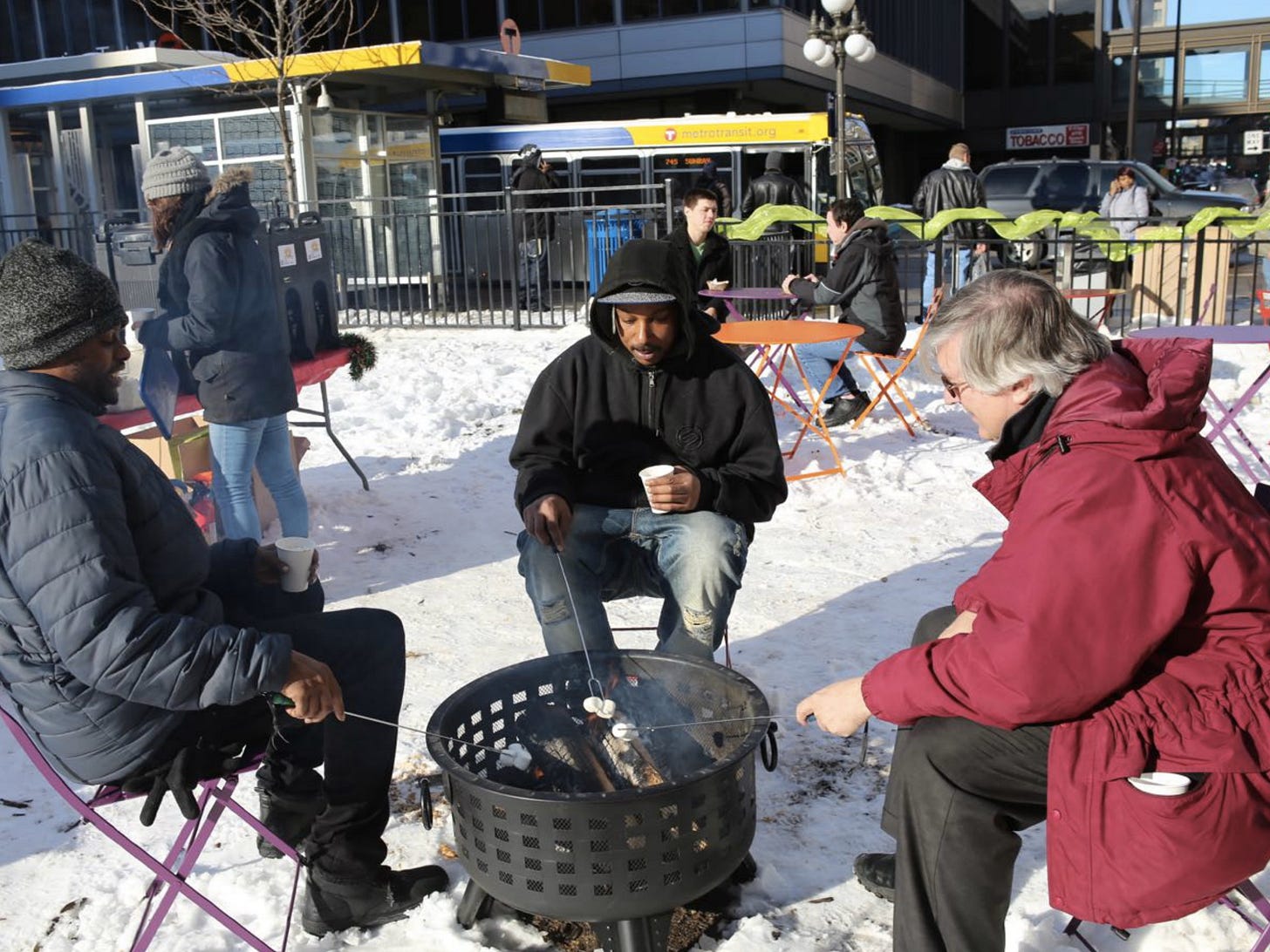 Three people sit around a fire pit roasting s'mores next to a transit stop in downtown St. Paul.