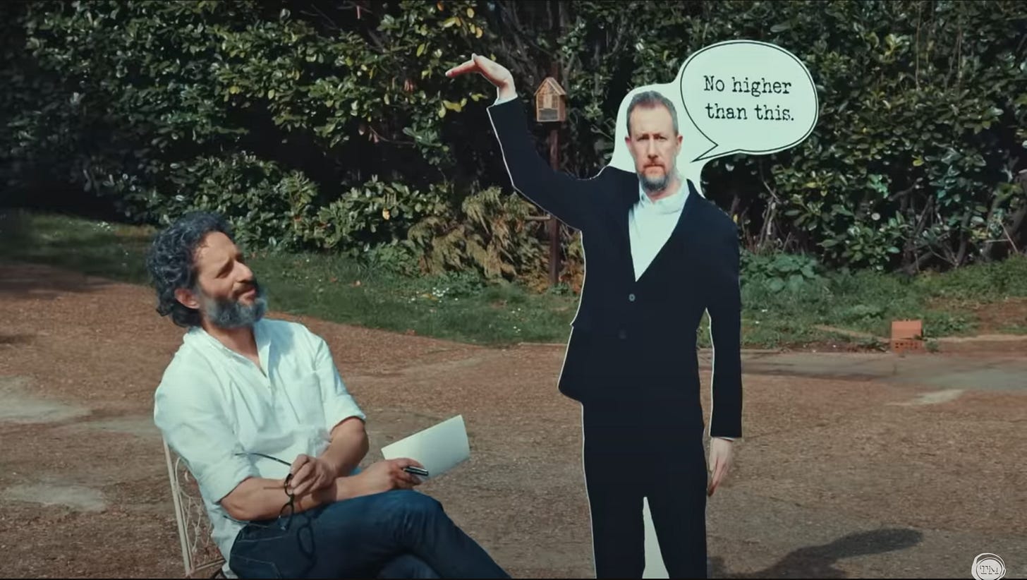 Jason Mantzoukas sits next to a life-sized cardboard cutout of Little Alex Horne.