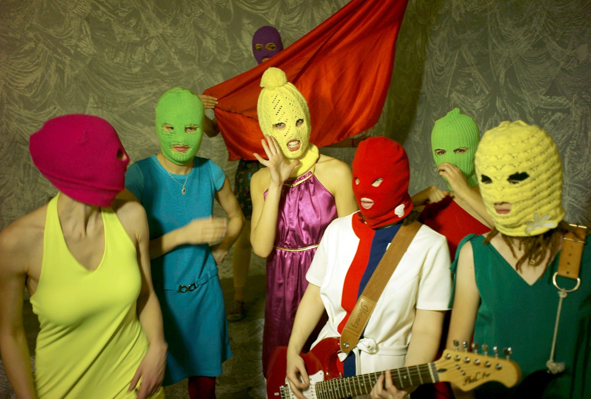 The pussy riot members wearing bright colored knitted masks. One member is holding a red flag and one is holding a guitar. They are all wearing bright dresses. The pussy riot members wearing bright colored knitted masks. One member is holding a red flag and one is holding a guitar. They are all wearing bright dresses.