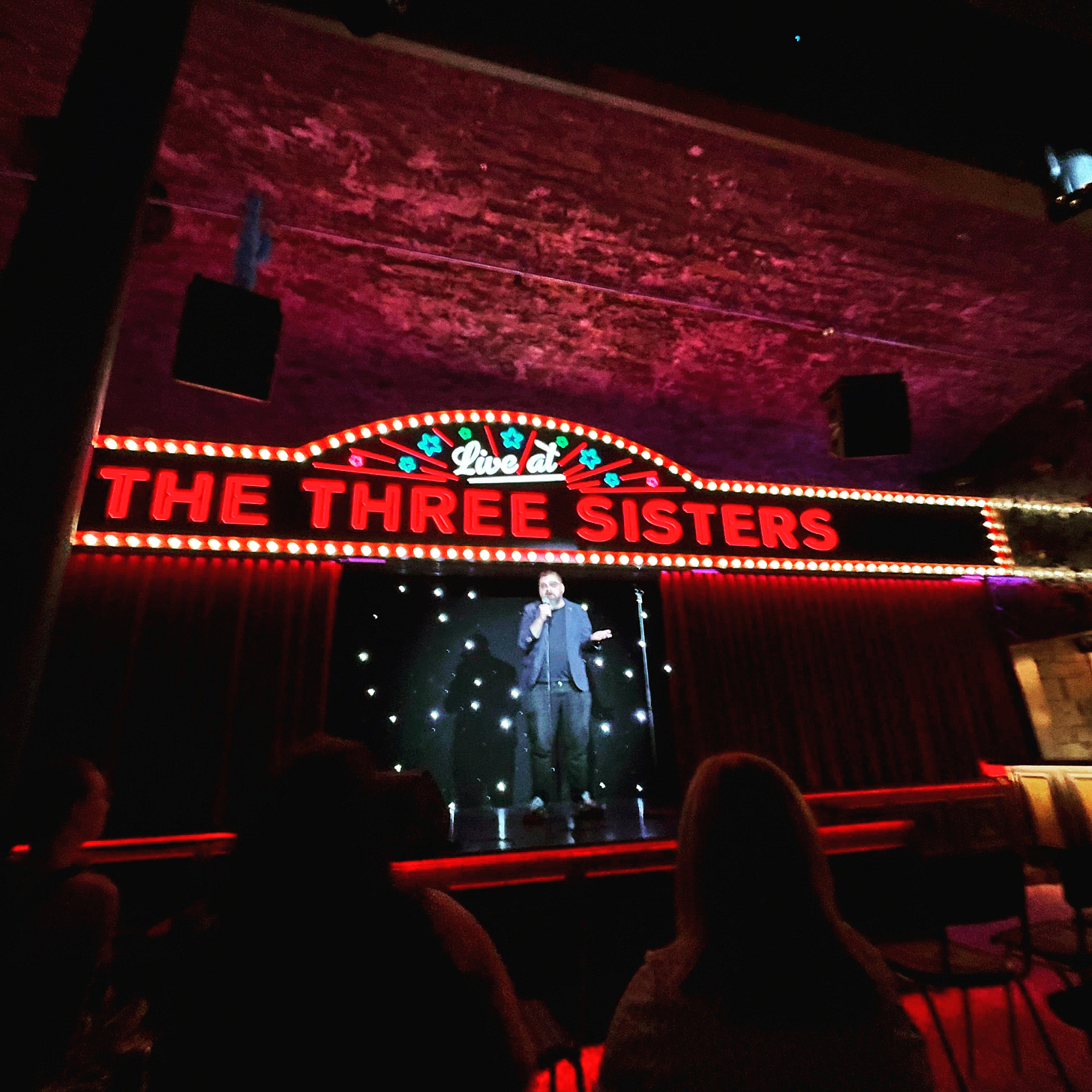 Me doing stand-up on a large raised stage, with a colourful electronic sign saying "Live at THE THREE SISTERS" above me