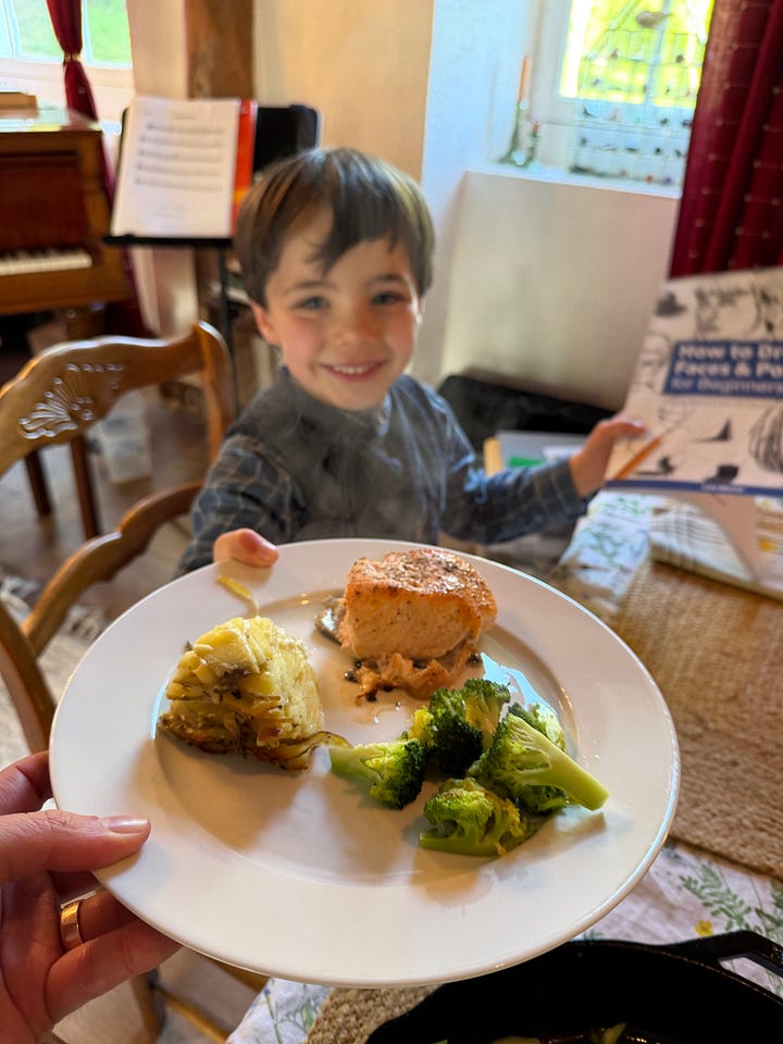 A boy at a table presenting a plate of salmon, potato and broccoli, and a bowl of homemade vanilla ice cream with chocolate chips.