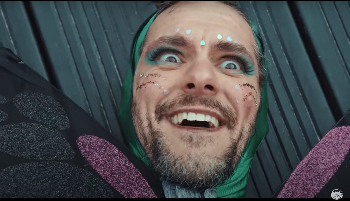 Mathew Baynton, dressed as a butterfly, wears glitter face paint and grins madly.