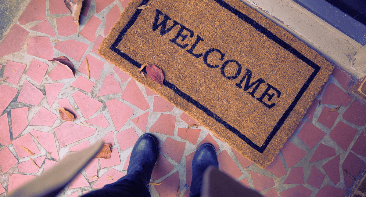 A person standing at the front door of a house, looking down onto a mat with 'Welcome' written on it A person standing at the front door of a house, looking down onto a mat with 'Welcome' written on it