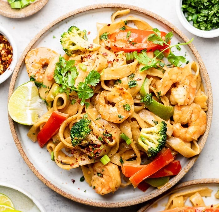 Plate of shrimp rice noodles with veggies and peanut sauce. It is topped with cilantro and has a lime wedge on the plate. Around it is another plate with shrimp noodles, small plate with lime wedges, bowl of red pepper flakes, bowl of chopped green onion, and a bowl of cilantro.