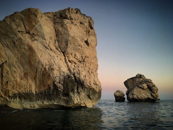 Rochedos no litoral de Pafos, no Chipre. Foto de Sergei Gussev/Flickr. Descrição de Imagem: Três rochedos se dividem na foto. À esquerda e bem perto, um rochedo enorme e cor de areia. Sua base está marcada por uma coloração mais escura e sua forma é mais regular. No meio e à direita, um rochedo irregular cor de areia. Ao fundo e bem menor do que os outros, mais centralizado, um pequeno rochedo arrendondado cor de areia. O céu não tem nuvens e o mar está calmo.