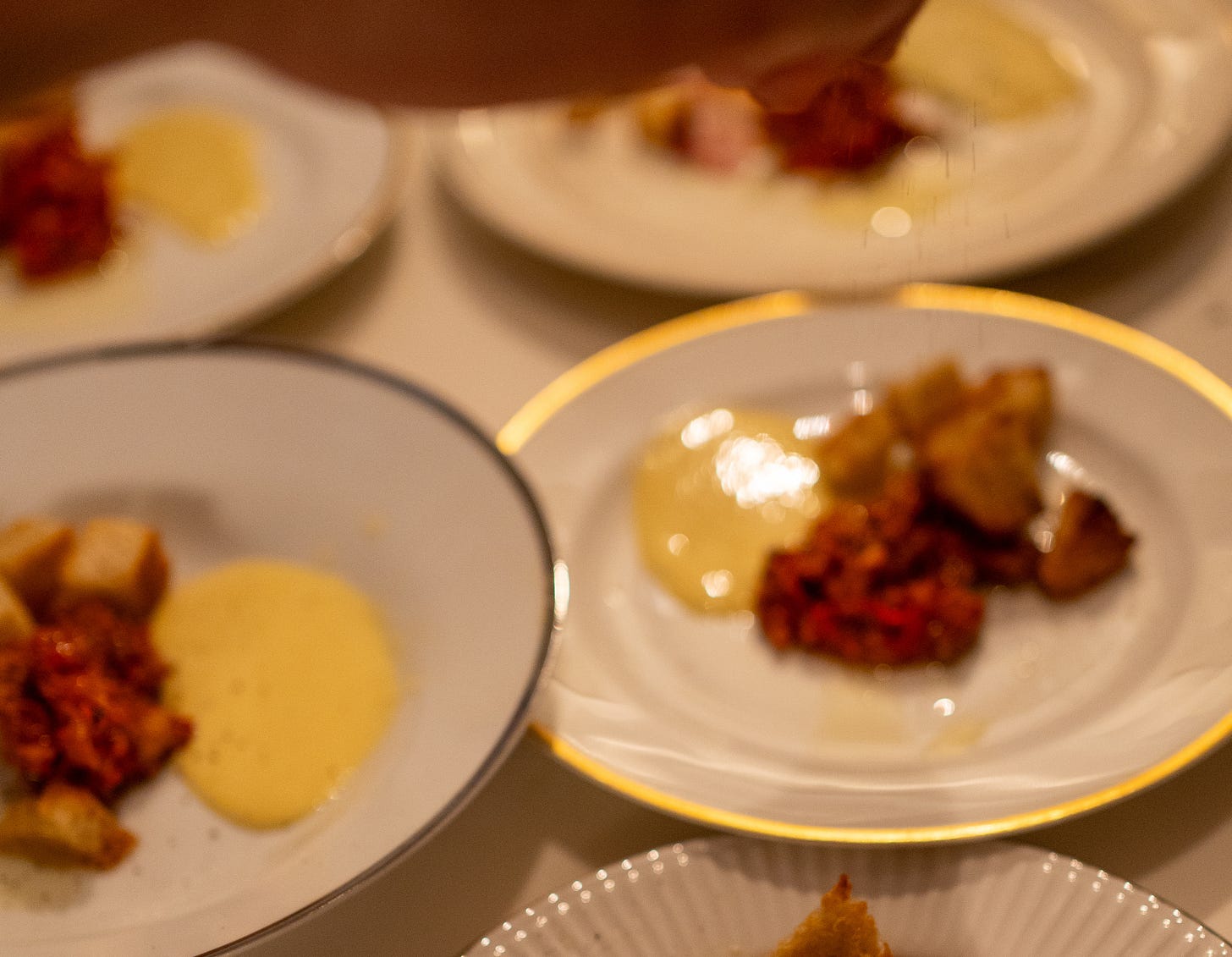 A hand reaching into frame to finish plating portions of heirloom tomato tartare, yellow aioli, and sourdough crisps on white plates with gold rims.