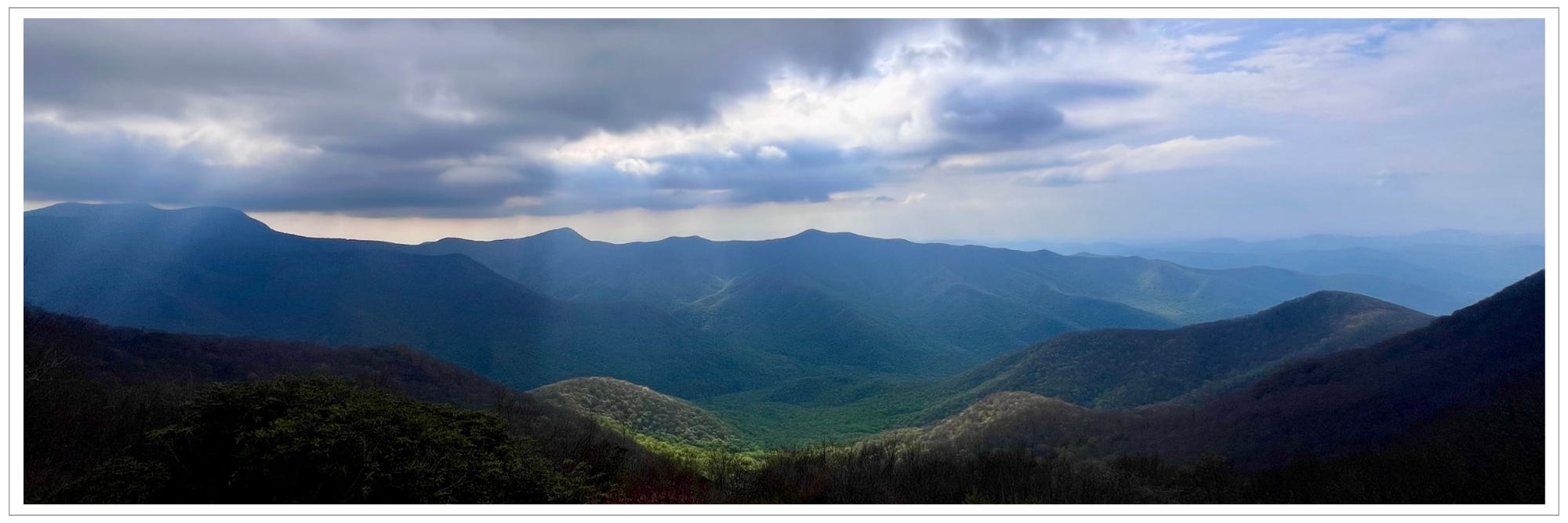 shafts of light breaking through holes in cloud cover, illuminating newly greening "bowl" in center of multiple mountain ranges