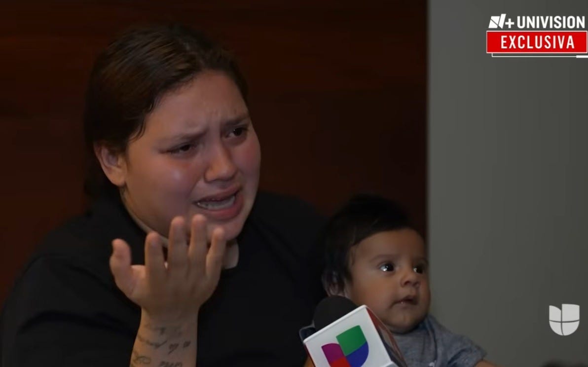 Video screenshot showing a distraught woman, gesturing as she weeps, sitting on a hotel bed holding her tiny two-month-old infant, who looks up with interest at the reporter (not on screen) interviewing the mother. The reporter's microphone, with Univision logo, is seen at the bottom of the shot. 