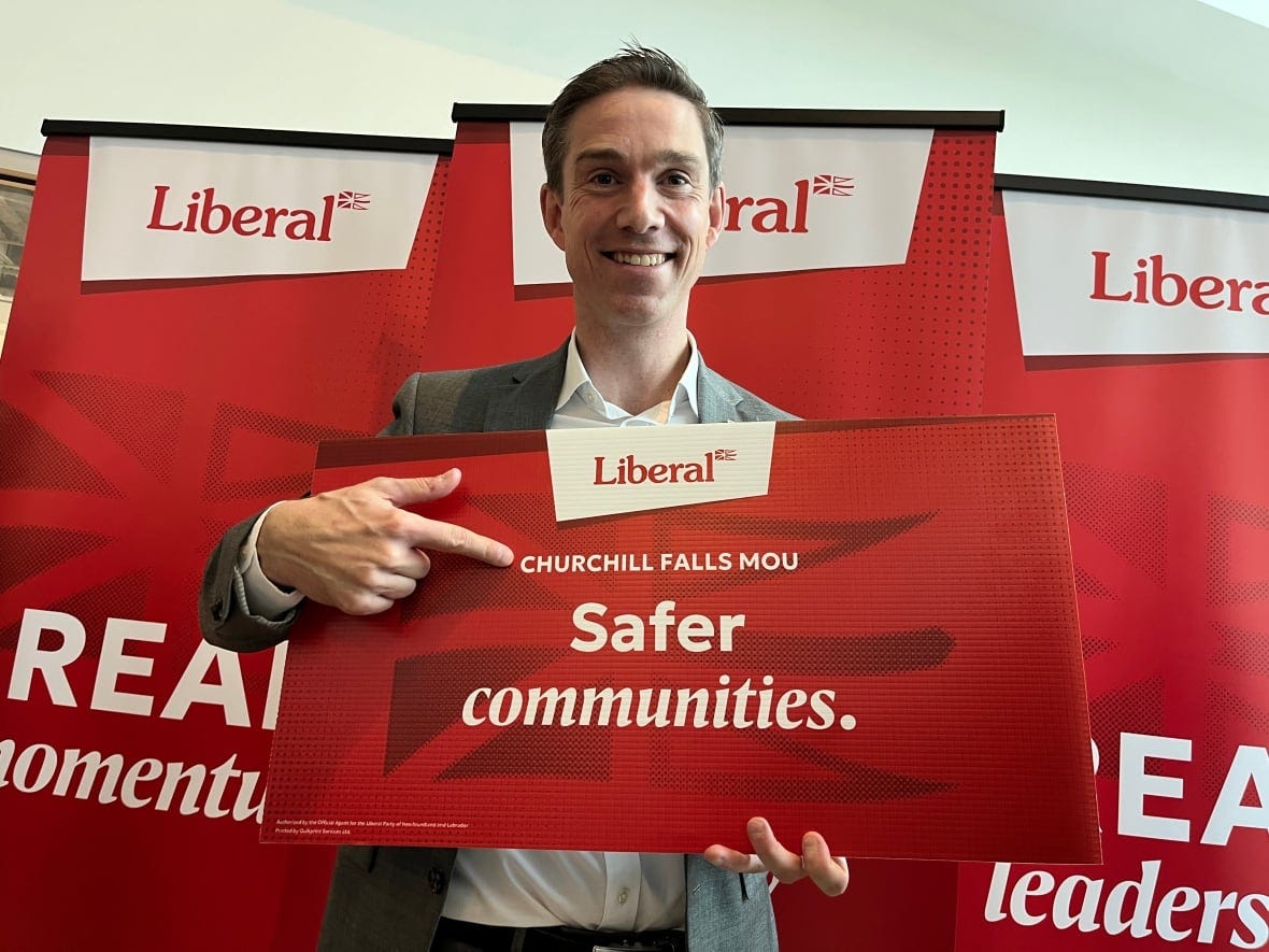 A smiling man holding a sign points to the sign's text. It reads 'Liberal,' 'Churchill Falls MOU' and 'Safer Communities.'