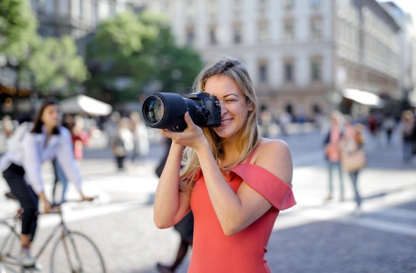 A smiling young woman with blonde hair wearing a red off-shoulder top takes a photograph with a professional DSLR camera on a busy city street. People and buildings are visible in the background, creating an urban atmosphere.