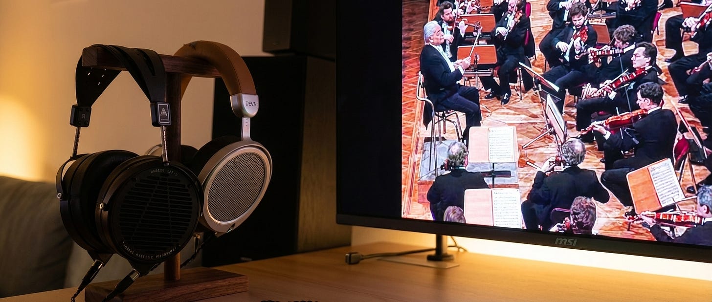 Two over-ear headphones on a wooden stand in front of a screen showing a classical orchestra and conductor.
