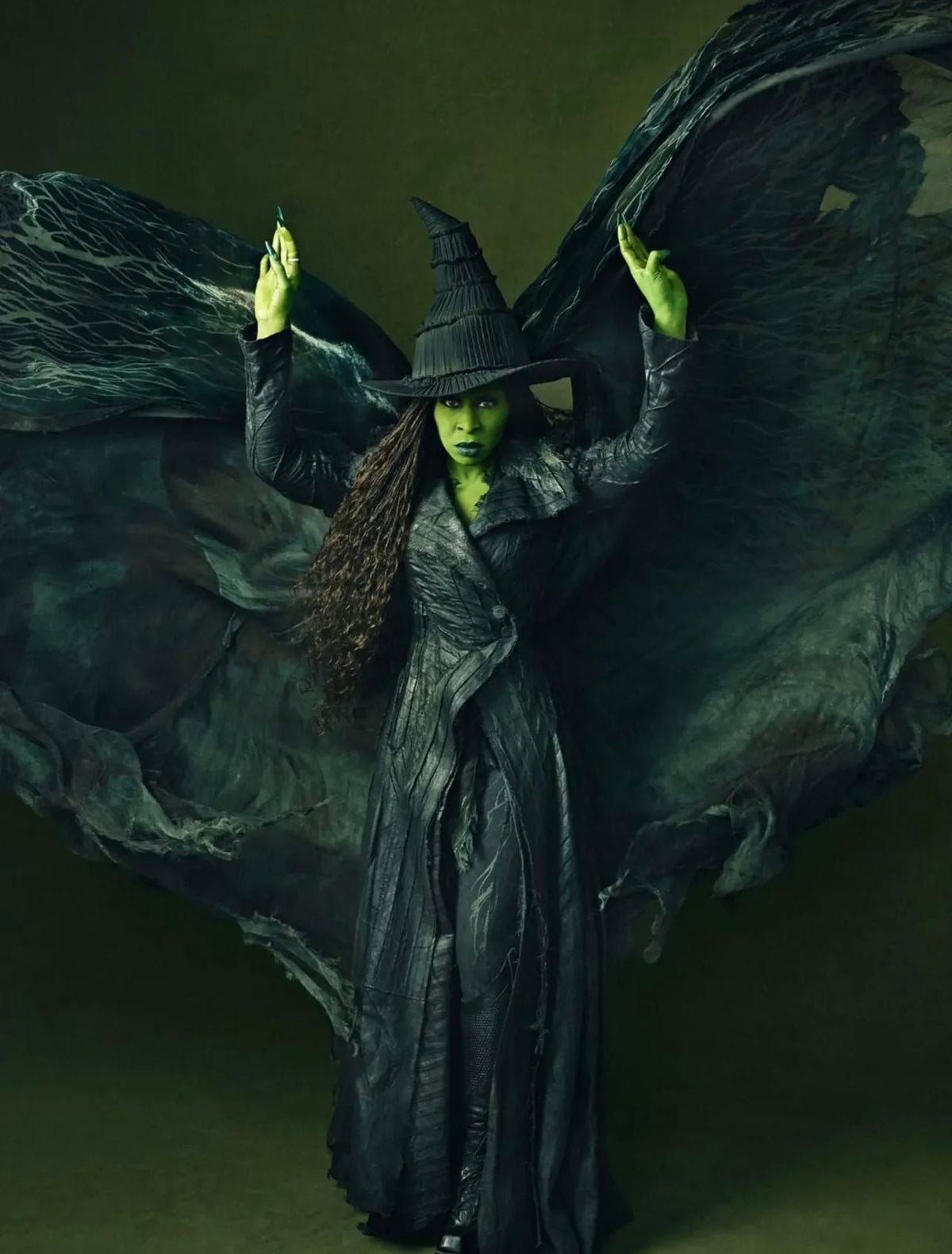 A person dressed as a dramatic witch stands with both arms raised, their skin painted bright green and long dark curls spilling from under a tall, twisted black hat. They wear a long, textured black coat that flares into torn, wing‑like panels of fabric spreading wide behind them, set against a dark green background.